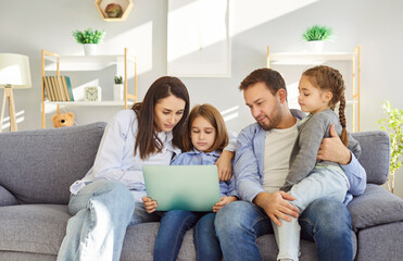 Young family of four sitting close together on sofa in living room, looking at laptop screen....
