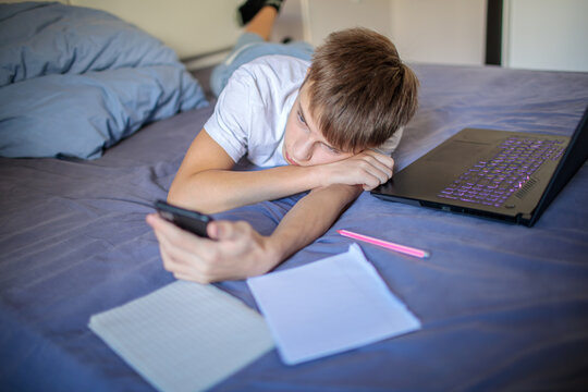 Bored teenage boy lying on his bed, procrastinating homework by scrolling on his smartphone next to a laptop and notebooks