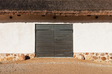 Old barn or countryside building with a wooden door. The wall is white plaster with a stone foundation and a thatched roof above. Traditional rural architecture detail