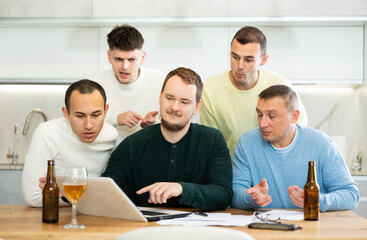 Group of men engaged in interested discussion with paperwork and laptop, accompanied by casual drinks at home kitchen..