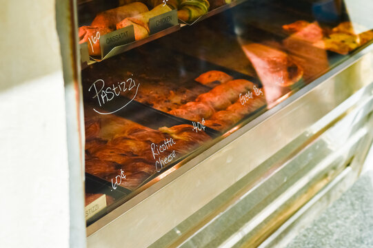 A view into a brightly lit glass display case of a traditional Maltese bakery, showcasing various types of freshly baked, savory Pastizzi. Handwritten labels indicate fillings like Ricotta Cheese.