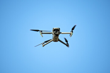 A drone is flying in the sky. The sky is blue and clear. The drone is in the air and is not touching the ground