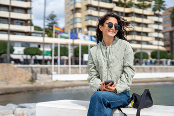 A stylish young woman with dark hair and sunglasses sits on a bench near the waterfront, holding her smartphone and smiling, with flags and modern buildings in the background