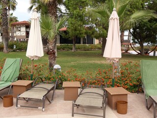 Tranquil poolside setup at ropical resort featuring beige umbrellas, sun loungers, wicker tables, and lush palm trees amid vibrant flower garden-perfect for travel, leisure, vacation lifestyle themes.