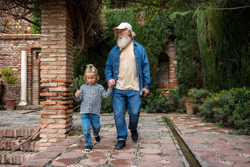 A grandfather and his grandson hold hands as they walk through a serene garden, surrounded by lush greenery and beautiful stone pathways