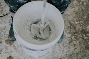 A young man mixes putty with a construction mixer in a bucket.