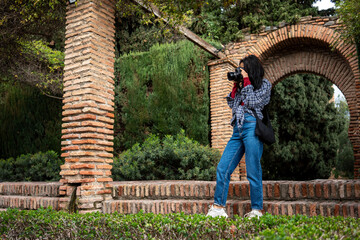 A woman dressed in a plaid shirt and denim jeans, holding a camera and photographing the surroundings in a lush garden. The architecture of the surrounding brick columns and greenery adds 