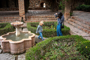 A child is playfully interacting with a fountain while a woman, likely a photographer, captures the moment. The child, wearing a plaid shirt, bends over to touch the water, while the photographer