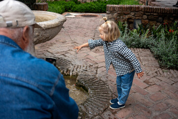 A young child playfully reaching for water at a fountain while an older man takes a photo