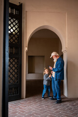 Elderly man and young child interact while looking at a smartphone inside a traditional architectural space with an arched doorway and a wooden lattice door