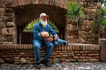 Elderly man and a young child sitting on a stone wall in front of an old brick wall, with a plant beside them, enjoying a peaceful moment together