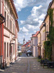 Beautiful historic street in Bielsko-Biała, Poland—charming old town architecture, cobblestone walkway, pastel facades, lush greenery, and a glimpse of church spire beneath a blue sky.​