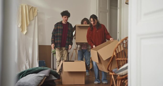 Friends carry large cardboard boxes into an apartment as they begin the process of moving house. Two guys and a girl bring in cardboard boxes