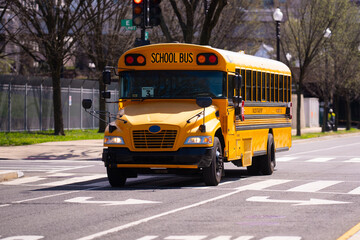 A yellow school bus drives through the city streets. The school bus provides safe transportation for students. Children commute daily on the school bus. The school bus stops at route.