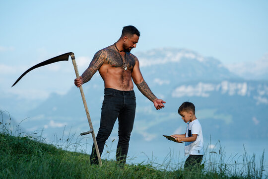 Farmers father and his son work together on farm field. The father teaches his child how to farm. Father and child on grass farm. The dad shows his son tablet. Son helps his father on farm.