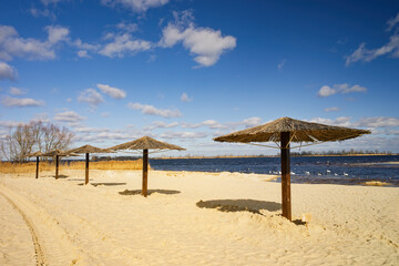 Straw parasols line a tranquil sandy beach on the Dnieper in Cherkasy, tire tracks in golden sand, swans on the wide river under a vivid blue sky with scattered clouds