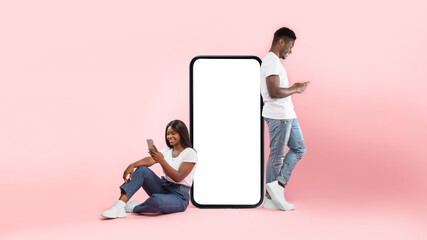 A young black couple interacts while leaning on a large blank smartphone screen. The woman smiles as she texts, and the man reads messages on his device, showcasing modern communication.