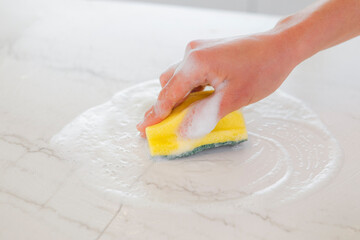 Hand using a sponge with suds to clean a white marble kitchen counter. Home hygiene and domestic work concept