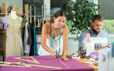 Focused male tailor sewing while young seamstress cuts textile using scissors in sewing workshop