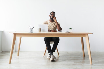 Fototapeta premium A black businessman enjoys a phone call while seated at his desk, surrounded by a laptop and notes. He shares a smile, indicating a positive conversation atmosphere in a bright studio setting.