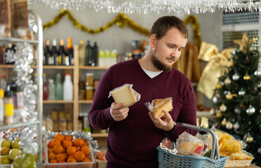 Young man chooses cheese in a supermarket with a Christmas tree in the background. Buying farm cheese at the festive grocery store.