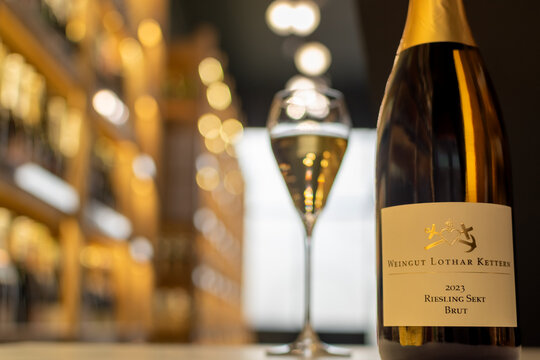 Bottle of Weingut Lothar Kettern Riesling Sekt Brut 2023 against blurred background with filled, sparkling wine glass and wine shelves in shop. Ukraine, Zhytomyr, October, 29, 2025