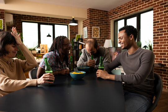 Men and women share laughter over funny joke, enjoying drinks and snacks in brick wall room. Cheerful group eats crisps and bursts into laughter as they trade amusing stories during cozy home hangout.