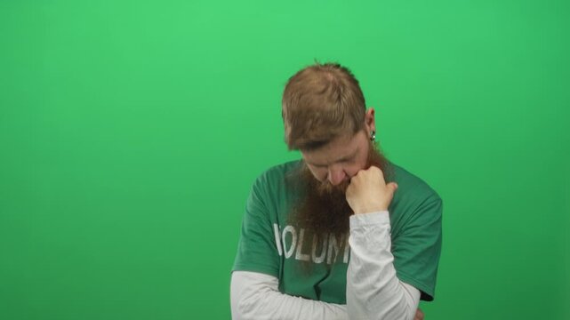 Young bearded volunteer man resting head on fist against green background in studio; pondering doubt.