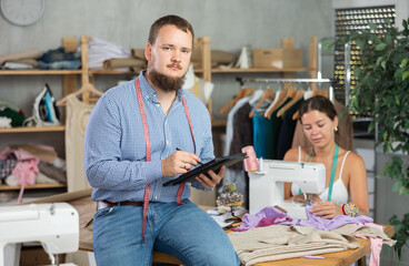 Young bearded tailor in checked shirt sitting on edge of worktable, sketching clothing designs on tablet with focused, creative look while female colleague working at sewing machine in cozy workshop