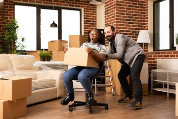 Young multiethnic couple enjoying playful moments in new home, laughing and riding on chair surrounded by cardboard boxes. Happy boyfriend and girlfriend celebrate moving day with fun and excitement.