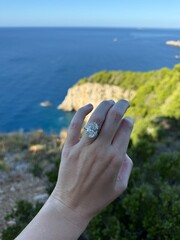 Vertical photo of a hand with an oval diamond engagement ring framed by the bright blue Adriatic Sea in Croatia, luxury travel jewelry content.