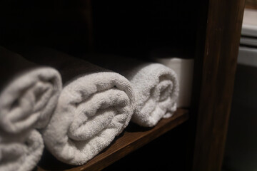 Rolled white towels neatly placed on a dark wooden shelf in soft light. Cozy minimalist composition...