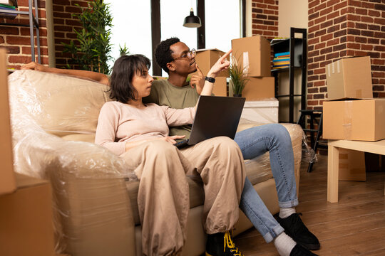 Husband and wife sit on unwrapped couch in living room, looking around while searching online for items to buy. Diverse couple with laptop, discussing furniture and decor ideas for new home.