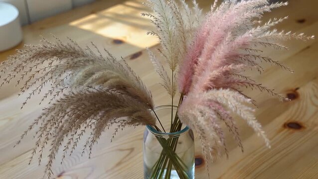 decorative arrangement featuring dried feathery plumes of ochre and pink colored pampas grass stems contained within a transparent glass vessel  resting atop a wooden table