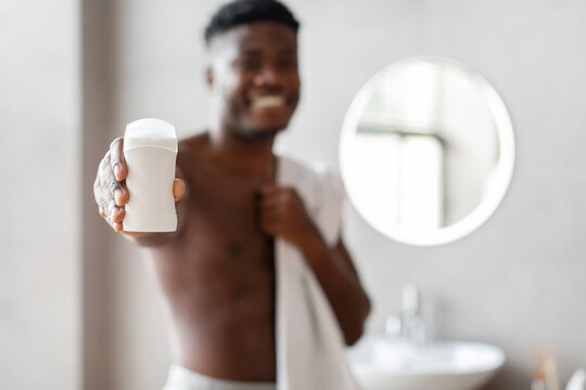 In a contemporary bathroom, a smiling African American man holds an antiperspirant stick while wrapped in a towel. He promotes male sweat protection and armpit hygiene at home.