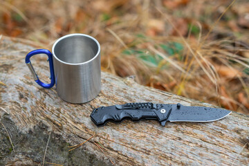 Outdoor camping gear on a rainy day with a folding knife and a metal mug on a wooden log
