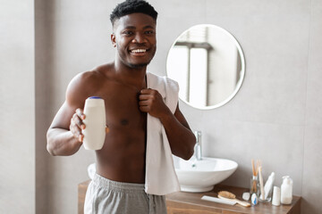 A smiling Black male stands in his bathroom, wrapped in a towel. He happily holds a bottle of...