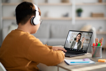 A overweight teen boy sits at a table, participating in a video conference with his tutor. He wears a headset and is focused on the laptop screen, engaging in learning from his home.