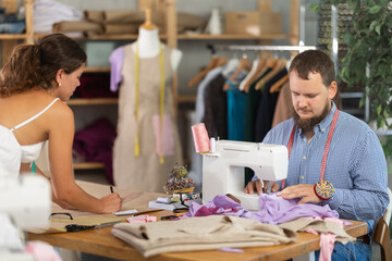 Focused male tailor sewing pastel fabric at machine with precision, while young female colleague sketching patterns on kraft paper nearby in shared studio