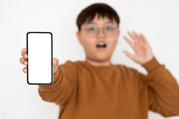 A shocked overweight Chinese teenager wearing glasses holds out a modern smartphone with an empty screen. He seems surprised while endorsing a fun mobile application in a blurred setting.