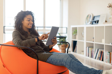 A young woman sits comfortably in an orange bean bag chair, using a tablet in her bright, modern...