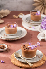 Three individual glasses of rich chocolate mousse, beautifully garnished with purple flowers and chocolate shavings, styled on a rustic surface.