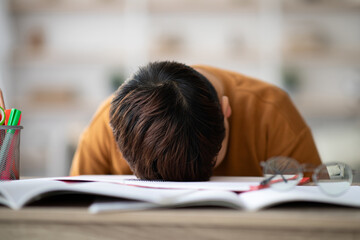 A chubby Asian boy lays his head on a desk covered with exercise books and stationery. He looks exhausted and bored while studying from home during the coronavirus pandemic.