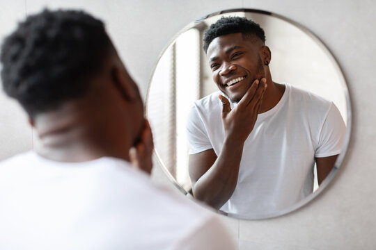 Black man stands near a round mirror in a modern bathroom, touching his unshaven chin while smiling at his reflection. He wears a simple t-shirt and seems to enjoy his grooming routine.