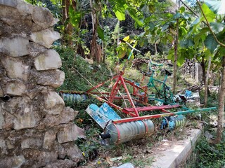 A rustic, possibly homemade, two-pontoon boat frame lies on a grassy, uneven bank amidst dense tropical trees and banana plants, with a simple shack visible in the background under an overcast sky.