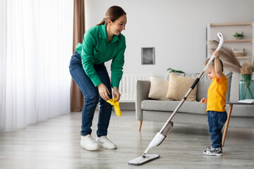 A mother and her young child are joyfully cleaning their living room. The mother holds a cleaning...