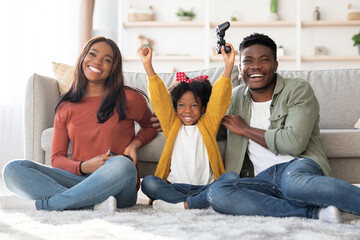 A family sits on a comfortable floor in their cozy living room. The child raises her arms in...