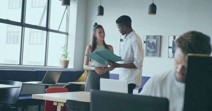 Discussing project coworkers reviewing green folder at laptop in open plan office, with lamps
