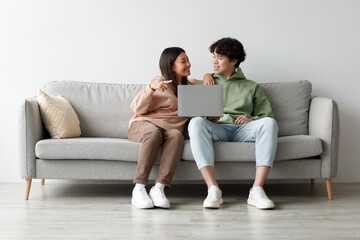Loving Asian couple sits together on a comfortable sofa at home, happily engaged in an online video...