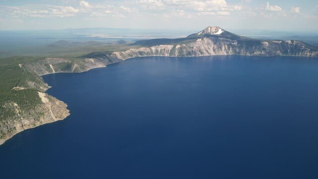 Aerial cinematic footage of Crater Lake National Park in Oregon, showcasing the vast caldera lake formed within an ancient volcano. The shot reveals the deep blue water surrounded by steep cliffs